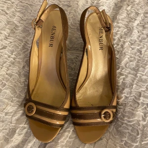 Bruna klackar från Menbur - Snygga bruna klackar från Menbur med öppen tå och dekorativa remmar med guldfärgade spännen. Perfekta för sommarens alla tillfällen.