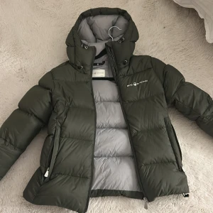 Grön dunjacka från Sail Racing - Säljer en grön dunjacka från Sail Racing i storlek XS. Jackan har en hög krage och huva för extra värme. Den är utrustad med dragkedja och har en stilren design. Perfekt för kyliga dagar.