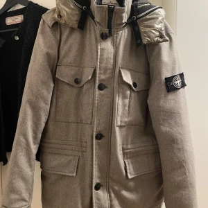 Ikoniskt Stone Island- jacka  - Snygg grå jacka från Stone Island med avtagbar huva i en kontrasterande beige nyans. Jackan har flera praktiska fickor med knappar och en stilren design. Perfekt för kyligare dagar med sin robusta look och ikoniska märke på ärmen. Jackan har vissa defekter på luvan (se bilder)