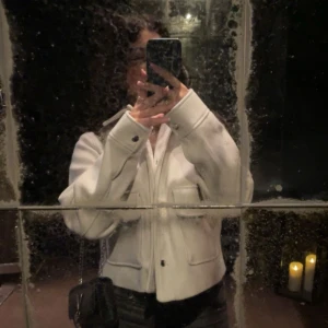 Vit jacka med fickor - Säljer en stilren vit jacka med knappar och stora fickor framtill. Jackan har en normal passform och långa ärmar, perfekt för en trendig look. Den är enkel att matcha med olika outfits.