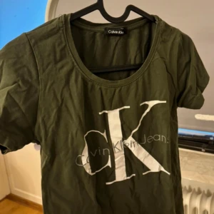 Grön t-shirt från Calvin Klein - Snygg grön t-shirt från Calvin Klein med stort tryck av CK-loggan på framsidan. Perfekt för en avslappnad stil. T-shirten har en rund halsringning och korta ärmar.
