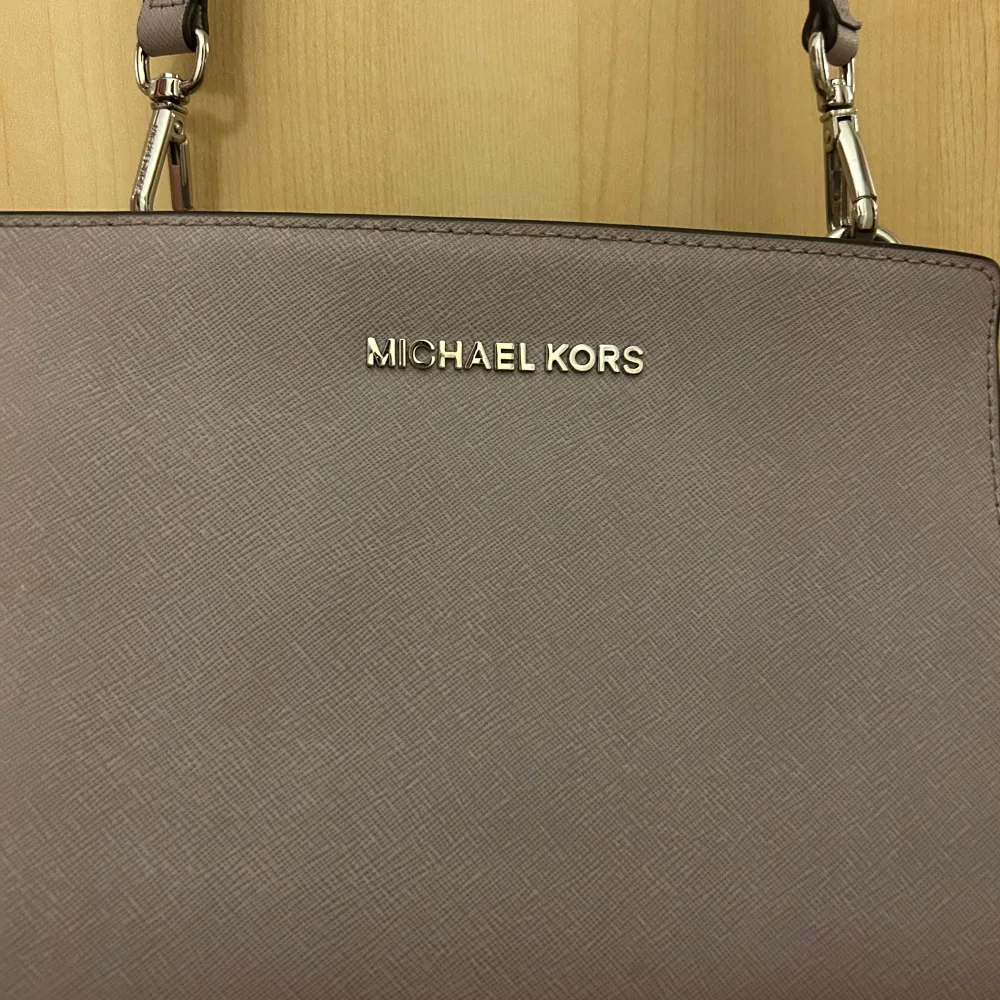 Snygg grå axelväska från Michael Kors med justerbar rem och dragkedja. Väskan har en stilren design med logotyp i metall på framsidan. Insidan är rymlig med flera fack för enkel organisering. Perfekt för att bära med sig det viktigaste med stil.. Laukut & Käsilaukut.