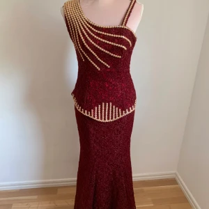 Vinröd glittrig klänning med pärldetaljer - Elegant vinröd klänning med glittrigt tyg och vackra pärldetaljer som pryder överdelen. Klänningen har en figurnära passform och är axelbandslös med en lång design som ger en sofistikerad look.