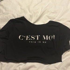 Svart croppad topp med text (This is me🇫🇷🇺🇸) - Säljer en svart croppad topp med texten 'C'EST MOI THIS IS ME' på framsidan. Perfekt för en avslappnad stil. Toppen har en lös passform och korta ärmar.