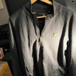 Svart cardigan från Lyle & Scott - Säljer en stilren svart cardigan från Lyle & Scott med dragkedja och broderad logga på bröstet. Perfekt för en casual look. Finns fler bilder vid förfrågan!