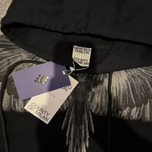 Svart hoodie från Marcelo Burlon - Snygg svart hoodie från Marcelo Burlon med ett unikt vingmönster i grått över axlarna och bröstet. Perfekt för en stilren look med en twist. Hoodien har en klassisk passform med långa ärmar och en bekväm huva. Storlek M och sitter bra på mig som är 184 och 75kg. Knappt andvänd och inga spår av andvändning!