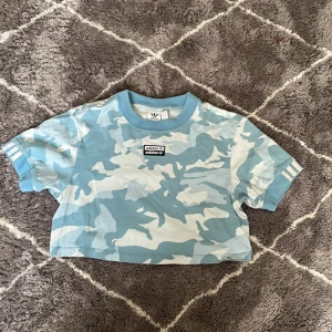 Blå camo croppad t-shirt från Adidas - Säljer en cool blå camo croppad t-shirt från Adidas. Den har korta ärmar med randiga detaljer och en logga på bröstet. Perfekt för en avslappnad stil!