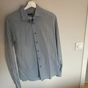 Randig skjorta från Riley - Snygg randig skjorta från Riley i blått och vitt. Skjortan har långa ärmar och knappar framtill. Perfekt för en stilren look.