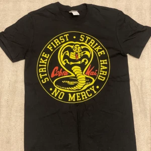 Svart Cobra Kai t-shirt från Gildan - Cool svart t-shirt från Gildan med Cobra Kai-tryck i gult och rött. Texten 'Strike First, Strike Hard, No Mercy' omger en orm. Perfekt för fans av serien! 👊🐍Aldrig använd. 