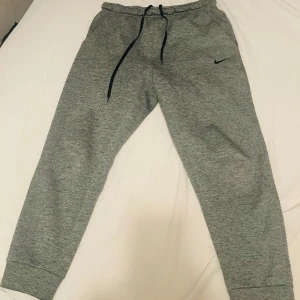 Grå Therma-Fit byxor från Nike - Säljer ett par grå mjukisbyxor från Nike med Therma-Fit teknologi. Byxorna har en justerbar dragsko i midjan och praktiska fickor och även en ficka med dragkedja. Perfekta för att hålla värmen under kyliga dagar.