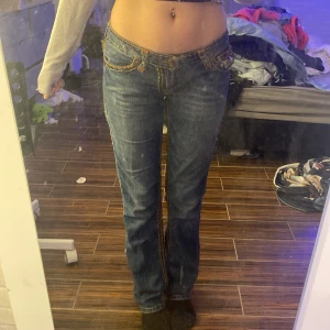 Blå jeans med broderade detaljer - Snygga blå jeans med broderade detaljer på bakfickorna och kontrastsömmar. De har en låg midja och en bootcut-stil som ger en avslappnad look. Perfekta för en casual outfit. Står storlek 29 US