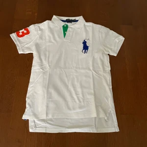 Vit kortärmad pikétröja från Ralph Lauren - Säljer en klassisk vit pikétröja från Ralph Lauren med korta ärmar. Tröjan har en broderad blå logga på bröstet och en orange siffra på ärmen. Den har en grön knappslå och är i custom fit.