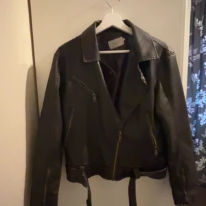 Svart skinnjacka från Nelly - Snygg svart skinnjacka från Nelly med klassisk bikerstil. Jackan har dragkedjor och ett bälte i midjan för extra edge. Perfekt för att ge din outfit en cool touch.