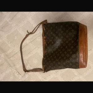 Louis Vuitton väska Noe - Säljer en snygg axelväska med klassiskt mönster och bruna läderdetaljer. Väskan har en justerbar axelrem och en praktisk dragskoöppning. Perfekt för dig som vill ha en stilren och tidlös accessoar.