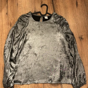 Silverfärgad blus med volangdetaljer - Snygg silverfärgad blus med volangdetaljer på axlarna. Blusen har långa ärmar och en liten öppning i ryggen. Perfekt för en glittrig look!
