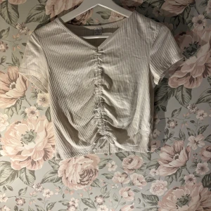 Beige ribbad topp med v-ringning - Säljer en beige ribbad topp med v-ringning och korta ärmar. Toppen har en snygg rynkad detalj framtill som ger en unik look. Perfekt för en stilren och modern outfit.