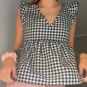 Svartvit rutig blus med volangärmar - Charmig svartvit rutig blus med volangärmar och öppen rygg. Blusen har en knytning i ryggen och en v-ringad halslinje. Perfekt för en somrig look!