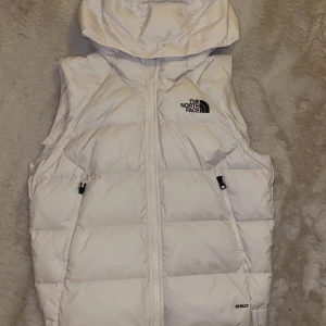Vit dunväst från The North Face - Säljer en snygg vit dunväst från The North Face med dragkedja och huva. Perfekt för kyliga dagar och har en stilren design med logotyp på bröstet.
