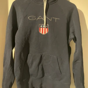 Mörkblå hoodie från GANT - Säljer en snygg mörkblå hoodie från GANT med ett broderat emblem och text på bröstet. Tröjan har en stor ficka framtill och justerbar huva med snören. Perfekt för en avslappnad stil.