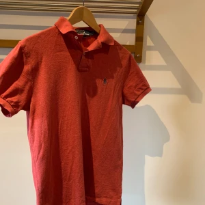 Röd pikétröja från Ralph Lauren (replika) - Snygg röd pikétröja från Ralph Lauren med klassisk krage och korta ärmar. Tröjan har en broderad logotyp på bröstet och är i en bekväm bomullskvalitet. Perfekt för en stilren look. ( replika)