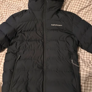 Svart dunjacka från Peak Performance - Säljer en svart dunjacka från Peak Performance med dragkedja och huva. Jackan har en stilren design med två praktiska fickor framtill. Perfekt för kyliga dagar. Väldigt bra skick.