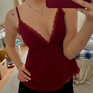 Vinröd spetskantad linne - Snyggt vinrött linne med spetsdetaljer och smala axelband. Toppen har en v-ringad design och elastisk midja för en bekväm passform. Oanvänd från Nelly.