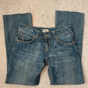 Low waist jeans - Snygga blå jeans från Hilfiger med klassisk femficksdesign och knappgylf. De har låg midja och är bootcut! Midjemåttet är 39cm rakt över och de går hela vägen ner till marken på mig som är 165cm