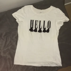 Vit t-shirt med texten HELLO - Säljer en vit t-shirt i bomull med texten 'HELLO' i svart och dekorativa tofsar under bokstäverna. Perfekt för en avslappnad stil.
