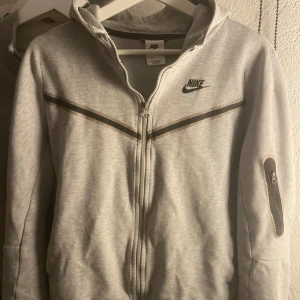 Grå huvtröja från Nike - Säljer en grå huvtröja från Nike med dragkedja och svart logga på bröstet. Tröjan är andvänd men i gott skick! Säljer den billigt för att den är för liten för mig. Hör av dig vid frågor!