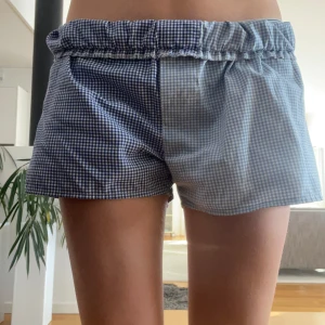 Rutig blå och vit shorts - Snygga och bekväma shorts med rutigt mönster i blått och vitt. De har en elastisk midja för en skön passform. Perfekta för en avslappnad dag hemma eller ute i solen.