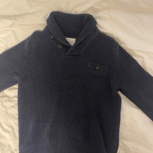 Mörkblå stickad tröja från NN.07 - Säljer en stilren mörkblå stickad tröja från NN.07. Tröjan har en unik krage med knappar och en dekorativ ficka på bröstet. Perfekt för kyliga dagar.