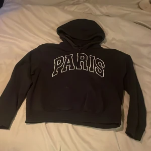 Mörkblå hoodie med Paris-tryck - Säljer en marinblå hoodie med texten 'PARIS' i vitt på framsidan. Tröjan har en klassisk huva och långa ärmar. Perfekt för en avslappnad stil.