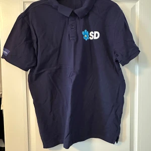 Mörkblå pikétröja med SD-logga - Säljer en mörkblå pikétröja med korta ärmar och SD-logga på bröstet. Tröjan har en klassisk krage med knappar och Sverigedemokraternas text på ärmen. Perfekt för dig som vill visa ditt stöd.