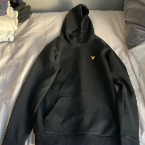 Svart hoodie från Lyle & Scott - Säljer en stilren svart hoodie från Lyle & Scott med en liten gul logga på bröstet. Perfekt för en avslappnad look med sin klassiska design och bekväma passform. Huvan har dragsko och tröjan har en praktisk magficka.