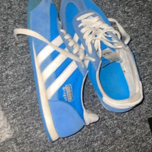 Blå Adidas dragon - Säljer ett par klassiska blå Adidas sneakers med vita ränder. Skorna har snörning och är i en sportig design, perfekt för en avslappnad stil.