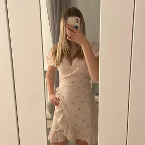 Ljusrosa omlottklänning från Gina Tricot - Charmig ljusrosa omlottklänning från Gina Tricot med små blommor och volangdetaljer. Klänningen har korta ärmar och ett knytband i midjan för en smickrande passform. Perfekt för en somrig look!