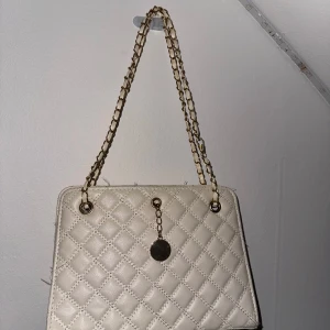 Beige quiltad handväska med kedja - Snygg vit handväska med quiltat mönster och guldfärgad kedja som axelrem. Väskan har en elegant design med en dekorativ guldfärgad detalj på framsidan. Perfekt för att ge din outfit en stilfull touch.