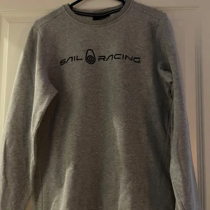 Grå tröja från Sail Racing - Säljer en stilren grå tröja från Sail Racing med svart logotyp på bröstet. Tröjan har långa ärmar och en rund halsringning. Perfekt för en avslappnad stil.