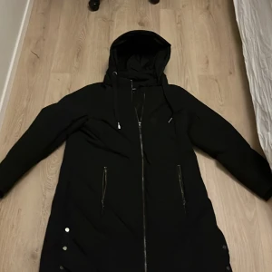 Svart parkajacka från Zizzi - Säljer en stilren svart parkajacka från Zizzi. Jackan har en praktisk huva och dragkedja framtill. Den är långärmad och har knappar som detaljer vid fickorna. Perfekt för kyliga dagar.