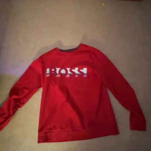 Röd tröja från Boss - Snygg röd tröja från Boss med vit logga på framsidan. Tröjan har långa ärmar och en klassisk passform. Perfekt för en stilren look.