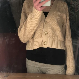 Beige stickad kofta med knappar - Säljer en beige stickad kofta med stora knappar framtill. Koftan har en croppad passform och långa ärmar med ribbade muddar. Perfekt för lager-på-lager-stil.