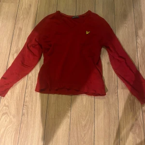 Röd tröja från Lyle & Scott - Snygg röd tröja från Lyle & Scott med v-ringning och broderad gul logotyp på bröstet. Perfekt för en stilren look.