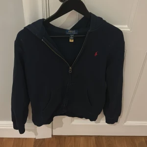 Mörkblå hoodie från Ralph Lauren - Säljer en snygg mörkblå hoodie från Ralph Lauren med dragkedja och röd logga på bröstet. 14-16 men går liten i storlek 