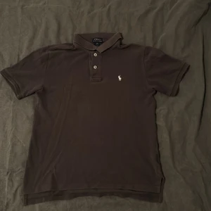 En mörkgrå pikétröja från Ralph Lauren - Säljer en väldigt mörk klassisk pikétröja från Ralph Lauren med den ikoniska loggan broderad på bröstet. Tröjan har korta ärmar och knappar vid kragen. Perfekt för en stilren och avslappnad look. Jag tar gärna byten.