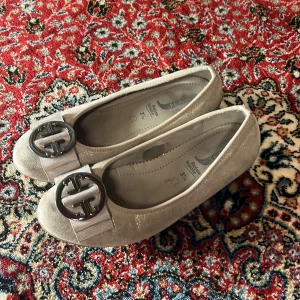 Beige ballerinaskor med dekorativt spänne - Snygga beige ballerinaskor från Jana med ett stort dekorativt spänne framtill. Skorna har en bekväm relax insole och är perfekta för en stilren look.