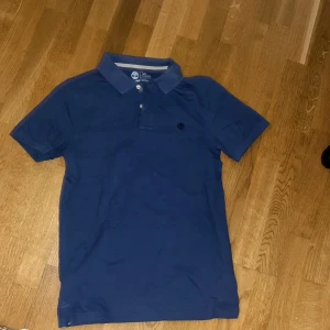 Blå pikétröja från Timberland - Säljer en snygg blå pikétröja från Timberland i 100% ekologisk bomull. Tröjan har en klassisk krage med knappar och en broderad logotyp på bröstet. Perfekt för en avslappnad stil.