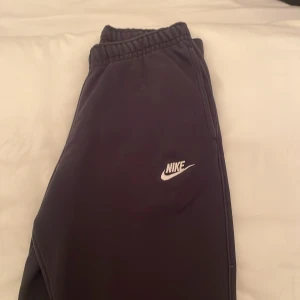 Svarta mjukisbyxor från Nike - Säljer ett par svarta mjukisbyxor från Nike med elastisk midja och snörning. Byxorna har en vit Nike-logga på benet och är perfekta för en avslappnad stil.