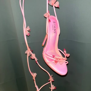 Rosa klackskor med fjärilar  - Säljer ett par rosa klackskor från Simmi. Skorna har en hög klack och knytning runt vristen för en feminin look. Perfekta för speciella tillfällen.