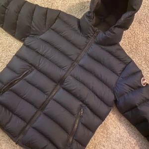 Marinblå pufferjacka från Colmar - Snygg marinblå pufferjacka från Colmar med huva och dragkedja. Perfekt för kyliga dagar med sin quiltade design och varma fyllning. Perfekt för en bakåtlutad still.