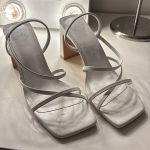 Vita sandaler med klack från H&M - Stilrena vita sandaler från H&M med smala remmar och fyrkantig tå. De har en elegant klack och är perfekta för sommarens festligheter. Sandalerna är tillverkade i syntetmaterial och har en modern design. Använda en gång 